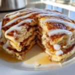 A stack of fluffy cinnamon roll pancakes, cut open to show the swirl layers, drizzled heavily with white icing.