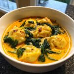 Close-up of a white bowl filled with creamy, orange coconut curry soup with dumplings and wilted spinach.