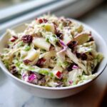 A close-up of a white bowl filled with creamy apple cranberry coleslaw, featuring visible chunks of apple and dried cranberries.