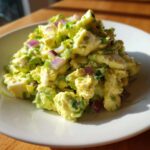 Close-up of creamy avocado egg salad mixed with chunks of hard-boiled egg, red onion, and green onion, served on a white plate.