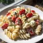 A close-up of creamy caprese pasta salad featuring fusilli pasta, cherry tomatoes, and small mozzarella balls coated in a creamy dressing and herbs.