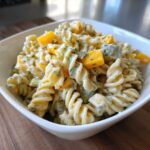 A close-up of creamy pickle pasta salad featuring rotini pasta, dill, and chunks of yellow bell pepper or pickle.