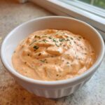 A close-up of thick, creamy, orange-hued remoulade sauce garnished with chopped parsley in a white ramekin.