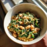 A close-up of creamy white beans with spinach and parmesan served in a white bowl, held up to natural light.