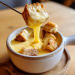 A piece of bread being dipped into a bowl of creamy, melted cheese fondue, showing a thick cheese pull.