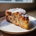 A moist slice of easy cinnamon apple cake, studded with apple pieces and dusted with powdered sugar, sitting on a white plate.