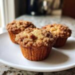Three freshly baked easy coffee cake muffins topped with a thick layer of cinnamon streusel, displayed on a white plate.