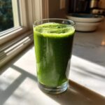 A tall glass filled with a vibrant, thick fat-burning green smoothie sitting on a sunlit marble countertop.