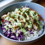 A close-up of a vibrant fish taco bowls featuring white rice, shredded cabbage, seasoned fish, and a generous drizzle of creamy green sauce.
