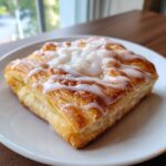 Close-up of a golden, flaky cheese danish topped with sweet white icing, resting on a white plate.
