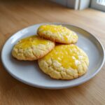 Three bright, crackled lemon sugar cookies topped with a shiny yellow lemon glaze, resting on a light grey plate.