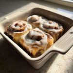 Four freshly baked gluten-free cinnamon rolls covered in thick white icing, sitting in a light-colored baking dish.