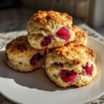 A stack of three golden-brown raspberry scones, bursting with bright red raspberries, sitting on a white plate.