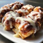 Close-up of several gooey cinnamon roll bites piled on a white plate, generously covered in thick white icing.