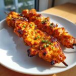 Two grilled bang bang chicken skewers coated in a vibrant orange sauce and topped with chopped green onions on a white plate.