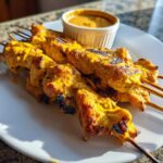 Close-up of four grilled chicken satay skewers with char marks, served on a white plate next to a small bowl of peanut sauce.