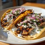 Two grilled mediterranean chicken tacos on pita bread topped with tzatziki, feta, and red onion.