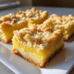 Four square slices of homemade lemon crumb bars featuring a bright yellow filling and a golden, sugary crumb topping.