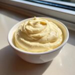 A white bowl filled with thick, pale yellow homemade mayonnaise, beautifully textured and catching sunlight.