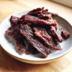 Close-up of a small bowl filled with strips of dark, glossy homemade teriyaki beef jerky sprinkled with spices.