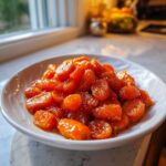 A white bowl filled with glistening, bright orange honey glazed carrots, set near a kitchen window.