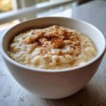 A close-up of creamy Jamaican green banana porridge topped generously with ground cinnamon in a white bowl.