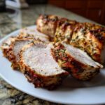 Close-up of sliced, juicy pork tenderloin recipe with a visible herb crust on a white plate.