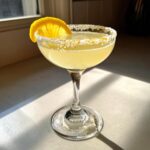 A bright yellow lemon drop martini in a coupe glass with a heavily sugared rim and an orange slice garnish.