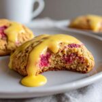 A close-up of a lemon raspberry cookie with a bite taken out, revealing bright pink raspberries and topped with thick lemon glaze.