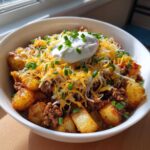 A close-up of a loaded potato taco bowl featuring seasoned potatoes, ground meat, shredded cheese, sour cream, and chives.