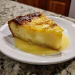 A moist slice of magic lemon cobbler served on a white plate with extra lemon sauce pooling around the base.