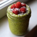 Close-up of vibrant green matcha chia pudding topped with fresh raspberries and shredded coconut in a glass jar.