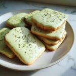 A stack of square matcha lavender sugar cookies topped with light green icing and sprinkled with lavender buds.