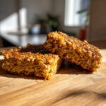 Two thick, moist squares of banana oatmeal bars resting on a wooden cutting board in bright sunlight.
