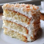 Close-up of a moist slice of three-layer coconut cake filled and frosted with white cream and topped with toasted coconut flakes.