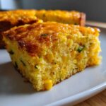 Close-up of a moist slice of Mexican cornbread, studded with whole corn kernels and green herbs, sitting on a white plate.