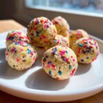 Close-up of several no bake cake batter protein balls rolled in colorful sprinkles, served on a white plate.