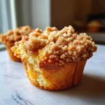 A close-up of a delicious peach cobbler muffin featuring a thick, sugary cinnamon crumb topping.