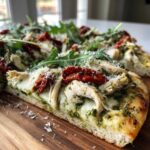 Close-up cross-section of a delicious pesto chicken flatbread topped with mozzarella, sun-dried tomatoes, and fresh arugula.