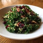A close-up of a vibrant quinoa kale salad mixed with pecans and dried cranberries served in a white bowl.