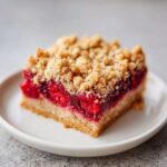 A close-up of one square raspberry crumble bars slice with a thick layer of bright red filling and buttery crumble topping.