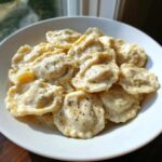 A white bowl filled with ravioli generously coated in a rich, creamy ravioli sauce and sprinkled with black pepper.