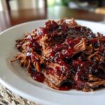 Close-up of tender, shredded slow cooker brisket generously coated in a thick, dark barbecue sauce on a white plate.