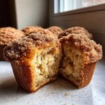 A close-up of a sour cream coffee cake muffin cut in half, showing the moist interior and thick cinnamon streusel topping.