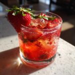 A close-up of a vibrant red strawberry basil smash cocktail filled with ice, muddled fruit, and garnished with a fresh strawberry.
