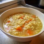 Close-up of a white bowl filled with hot pastina soup, featuring small star-shaped pasta, sliced carrots, and herbs in a clear broth.