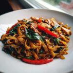 A close-up, appetizing photo of freshly made thai drunken noodles mixed with chicken, basil, and bright red chili peppers.