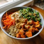 A close-up of a vibrant thai peanut sweet potato buddha bowl featuring roasted sweet potatoes, shredded carrots, quinoa, and cilantro, drizzled with peanut sauce.