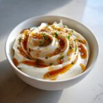 A close-up of a creamy, beautifully swirled whipped ricotta dip topped with honey, chili flakes, and chopped chives.