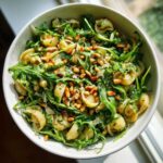 Overhead view of a white bowl filled with zesty lemon arugula pasta salad, topped generously with toasted pine nuts.